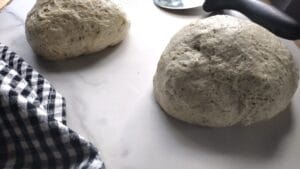 2 homemade pizza dough balls with a black checkered towel and pizza cutter along side