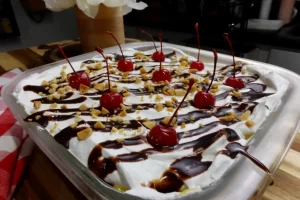 Whole pan of No-Bake Banana Split Dessert layered with bananas, vanilla pudding and whipped topping, topped with chocolate syrup, peanuts and cherries.
