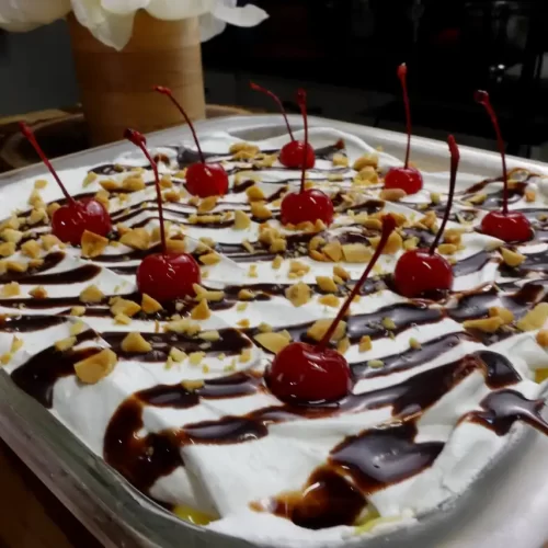 Whole pan of No-Bake Banana Split Dessert layered with bananas, vanilla pudding and whipped topping, topped with chocolate syrup, peanuts and cherries.